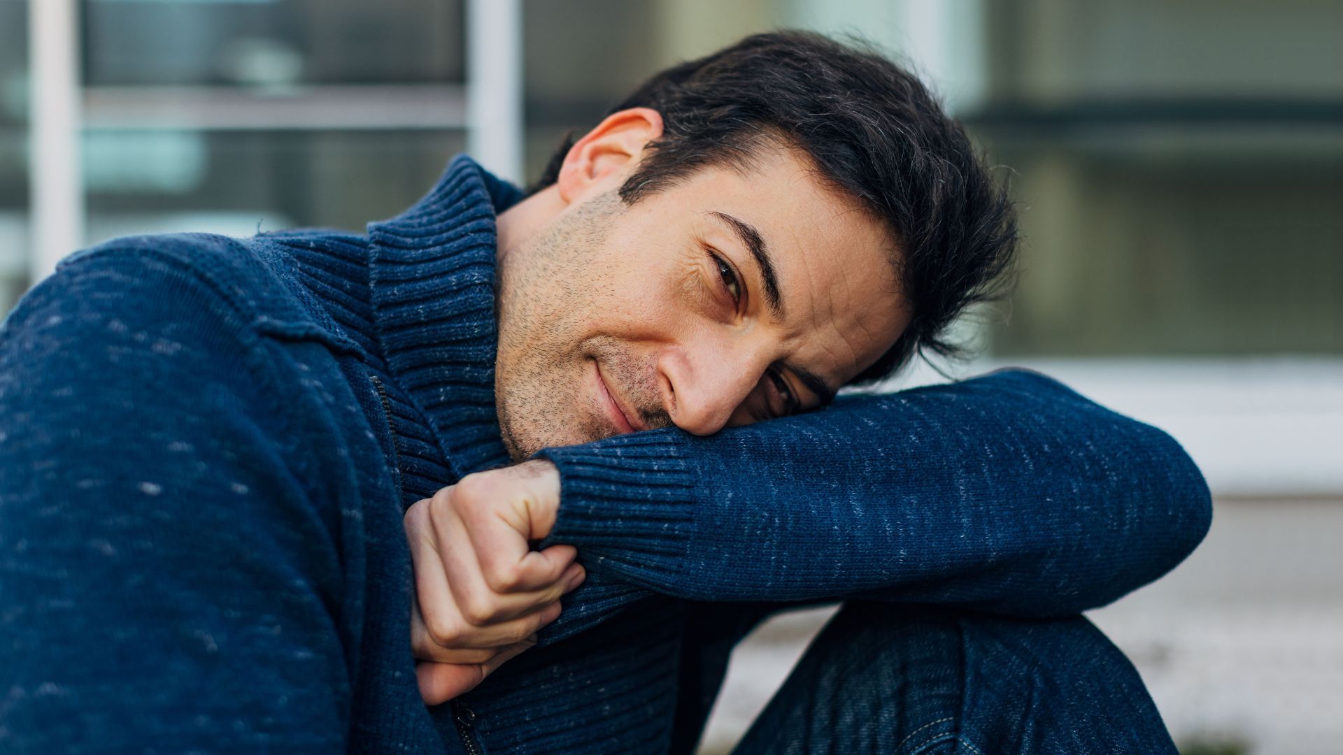 Homme en pull bleu reposant son menton sur son bras, souriant doucement.